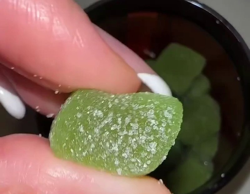 Des doigts tiennent un gummy vert au-dessus du pot
